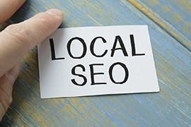 A close-up of a hand holding a paper with the written text local SEO on it against a wooden teal-colored surface.